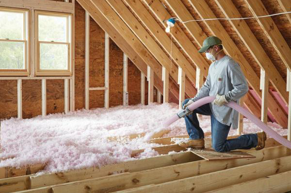A worker in a mask is blowing insulation into an unfinished attic with exposed studs